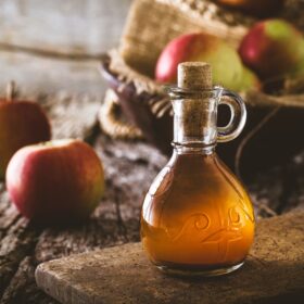 Decorative glass bottle of amber-colored apple cider vinegar with cork stopper on rustic wooden surface, fresh apples in background, natural wellness composition