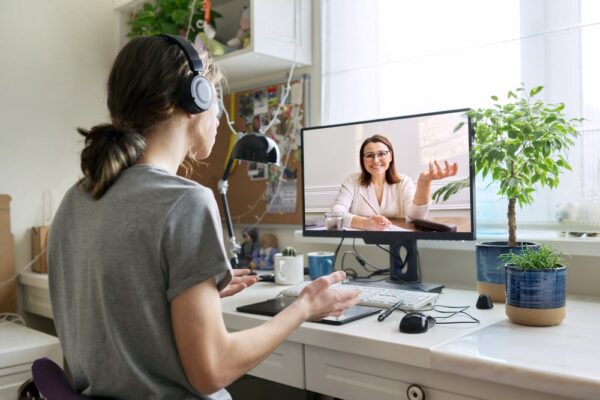 Person wearing headphones having video call with counselor on computer monitor, sitting at home office desk with plants and personal items in background