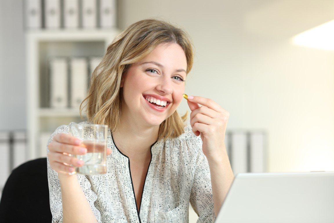 Woman in white patterned blouse smiling while taking supplement with glass of water at desk, laptop visible in bright office setting