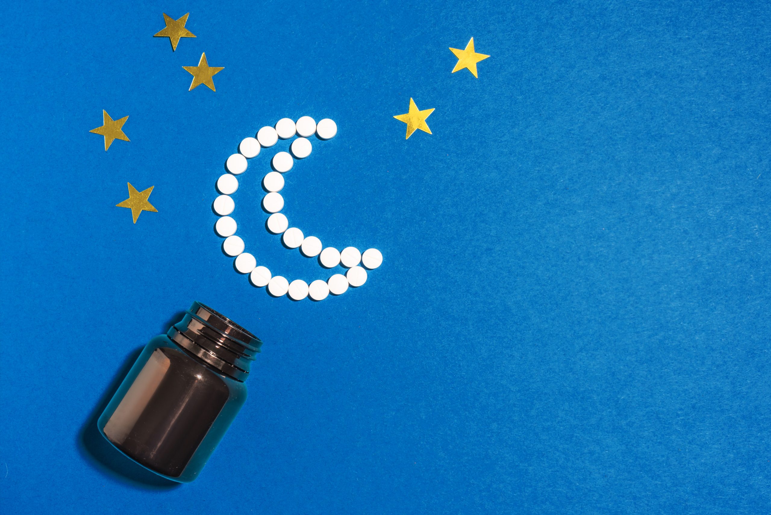 Melatonin tablets in shape of crescent moon on dark blue background, copy space. Bottle of dietary supplement for healthy sleeping. Insomnia concept.