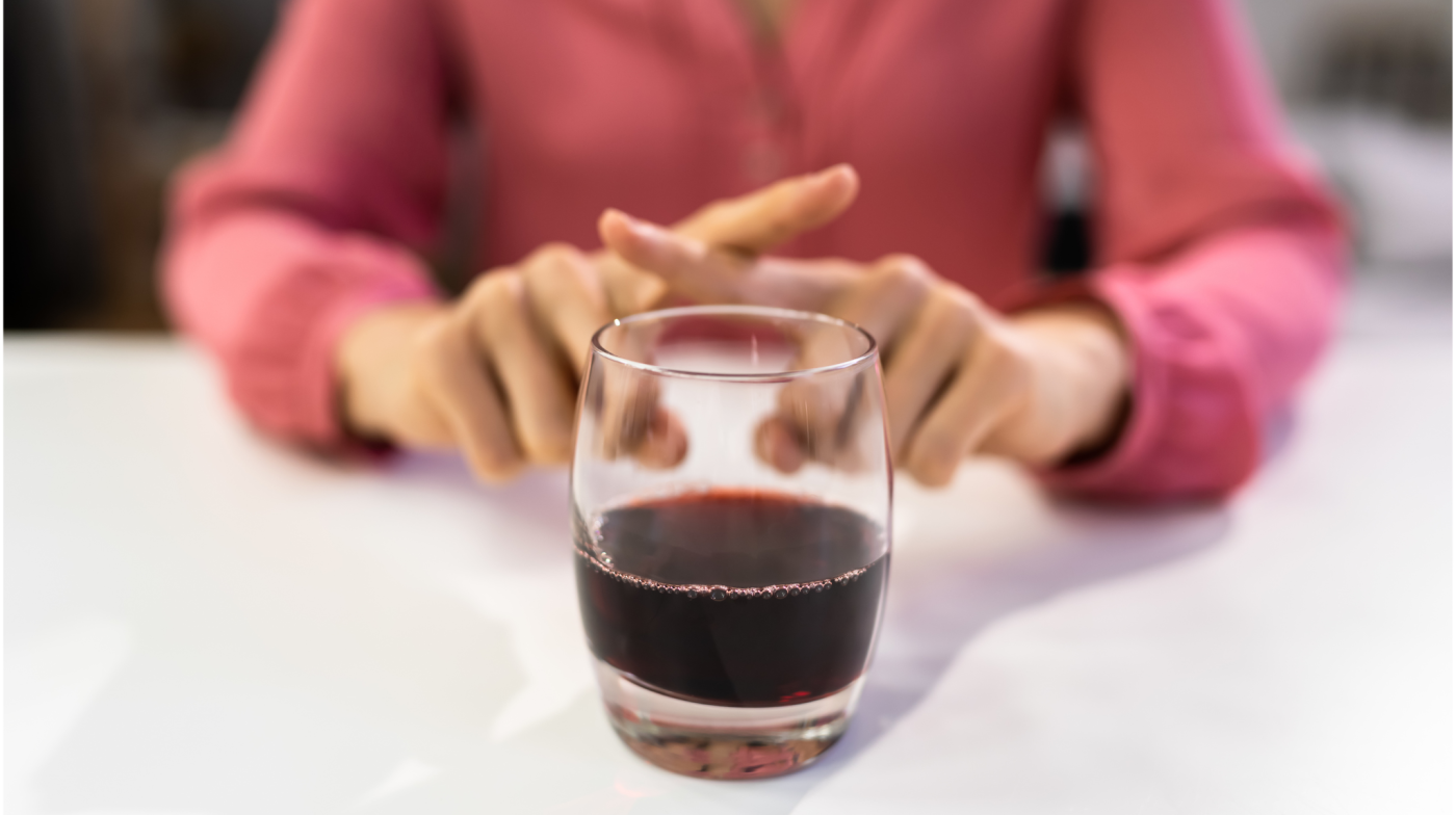 Woman in a pink shirt with her fingers in an x behind a glass of red wine to suggest saying no to drinking alcohol